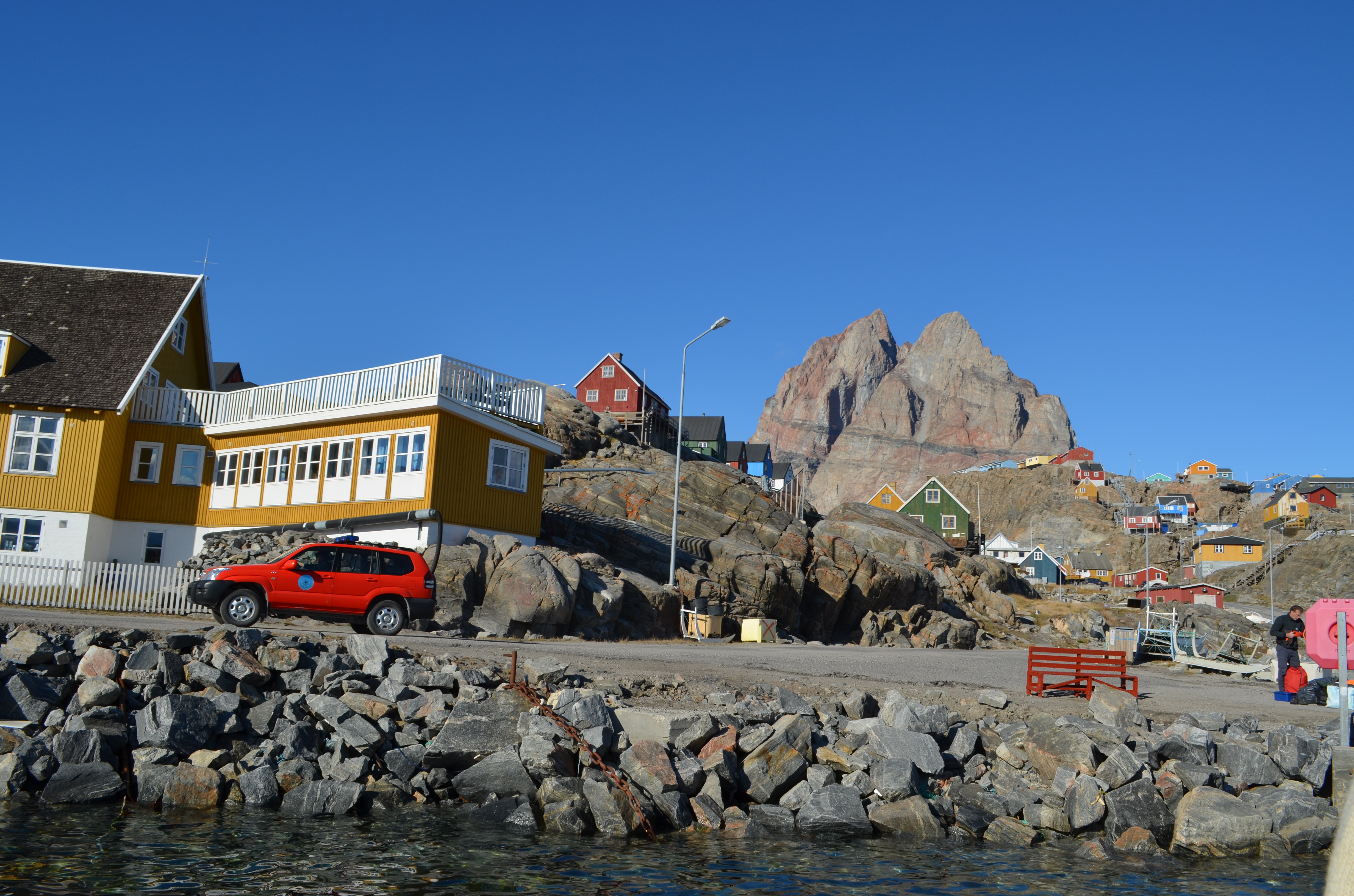 Spring landscape in Greenland, coloured houses along the coastline, and a tall mountain in the backgorund. 